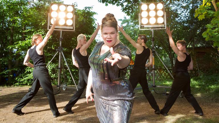 Dani Paschulke tanzt und singt im Bühnen-Outfit zusammen mit Tänzern vor dem Bauwagen