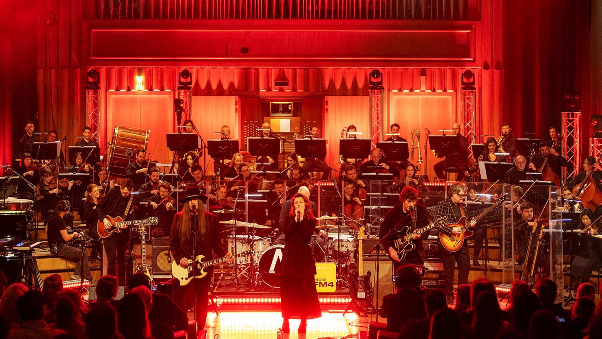 Man sieht einen beleuchteten Konzertsaal mit einer Band, einer Sängerin, einem Orchester und Publikum.