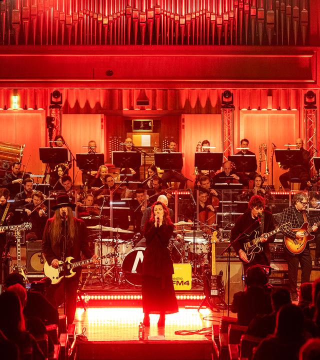 Man sieht einen beleuchteten Konzertsaal mit einer Band, einer Sängerin, einem Orchester und Publikum.