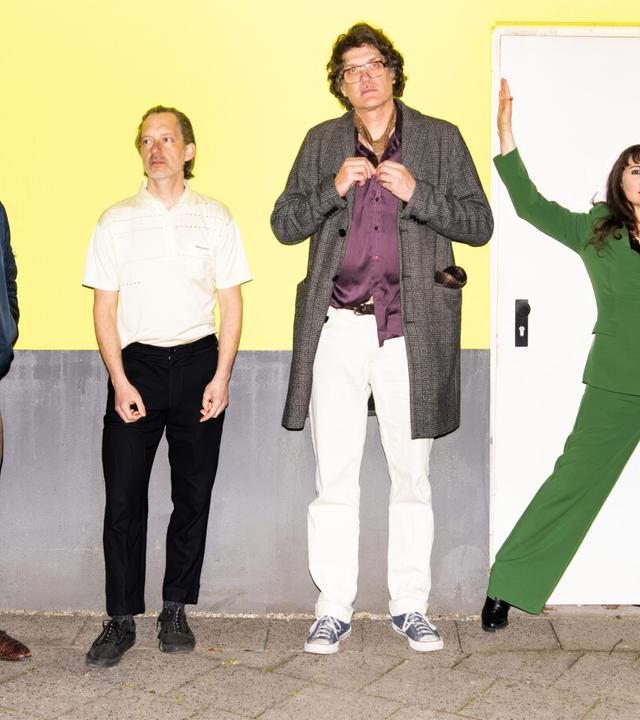 Drei Männer und eine Frau stehen vor einer gelb-grauen Wand.