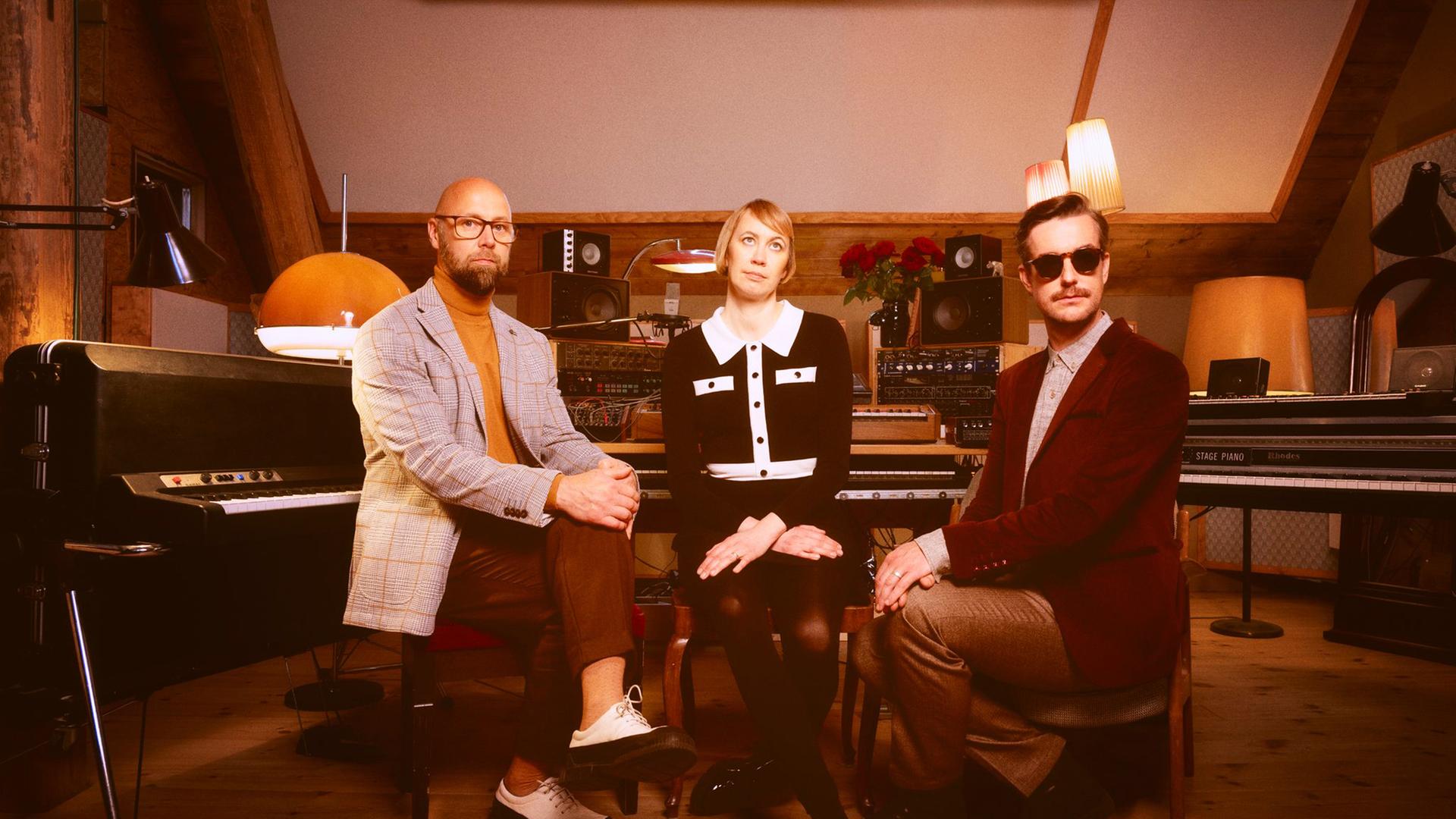 Zwei Männer und eine Frau sitzen in einem Musikstudio im Stil der 1970er Jahre.