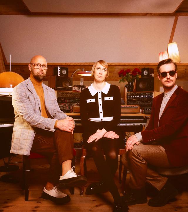 Zwei Männer und eine Frau sitzen in einem Musikstudio im Stil der 1970er Jahre.