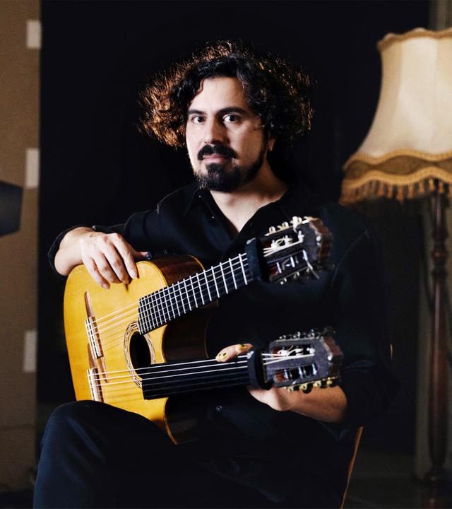 Ein Mann mit einer Doppelhals- Gitarre sitzt zwischen einem Scheinwerfer und einer Stehlampe.