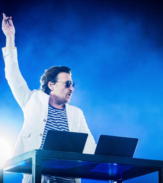 Ein Mann, der einen weißen Anzug trägt, streckt den Arm empor. Er steht hinter einem DJ-Pult auf einer Bühne.