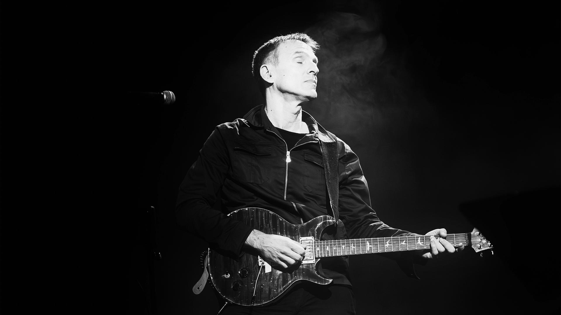 Man sieht eine Schwarz-Weiß-Fotografie auf der ein Mann zu sehen ist, der E-Gitarre spielt.