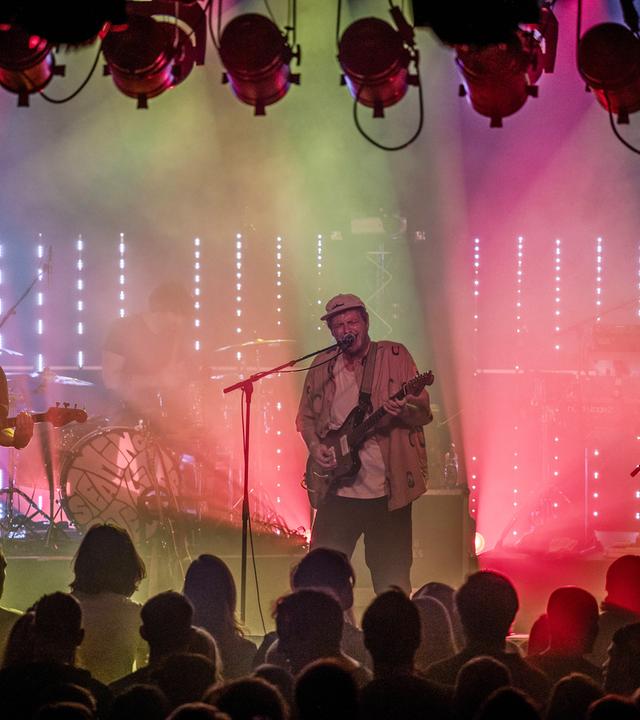Eine Rockband spielt auf einer Bühne, die von grünen und lilafarbenen Lichtern angestrahlt wird. Im Vordergrund steht das Publikum.