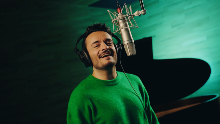 Giovanni Zarrella steht singend, in einem grünen Pullover vor einem Mikrofon, das von der Decke hängt. Im Hintergrund ist ein aufgeklappter Flügel.