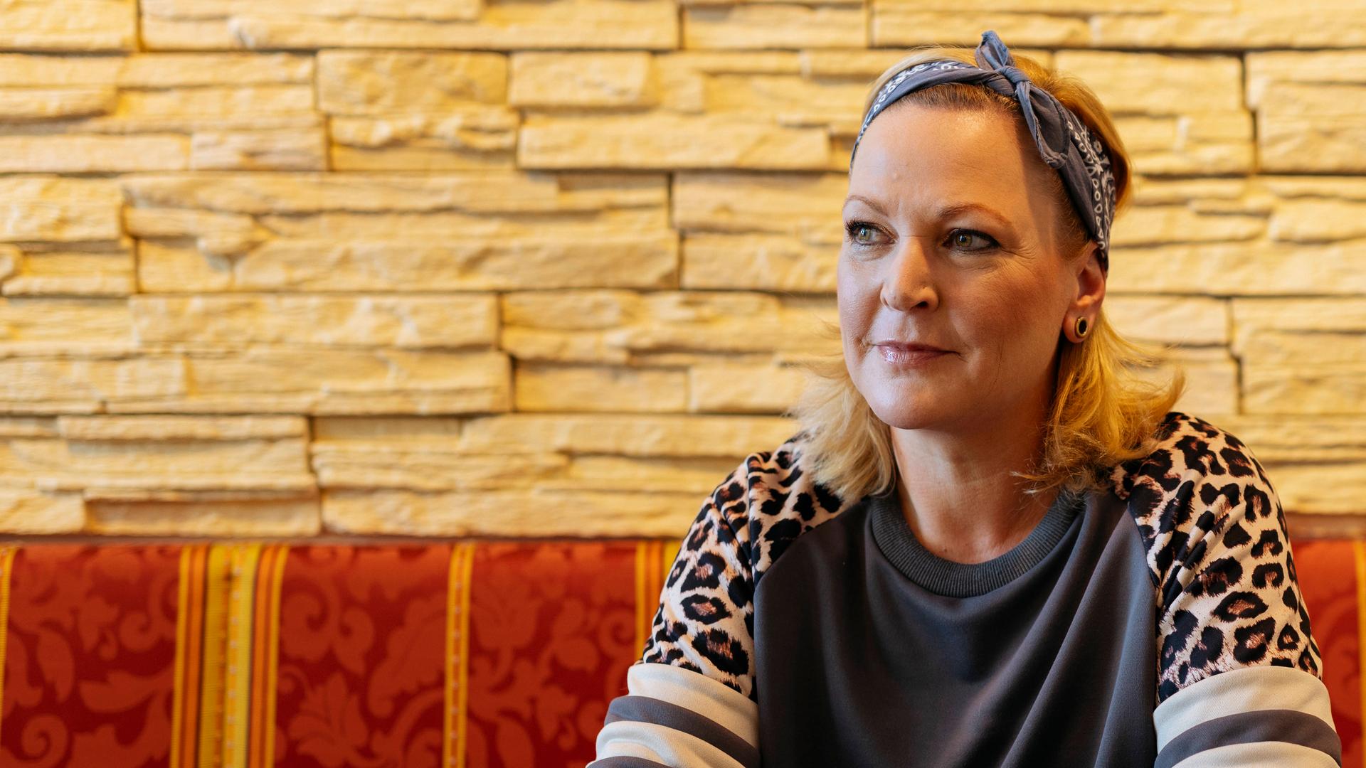 Martina sitzt auf einer Bank, an einem Tisch und schaut zur Seite. Sie hat die Hände auf den Tisch gelegt und verschränkt. Vor ihr steht eine weiße Tasse. 