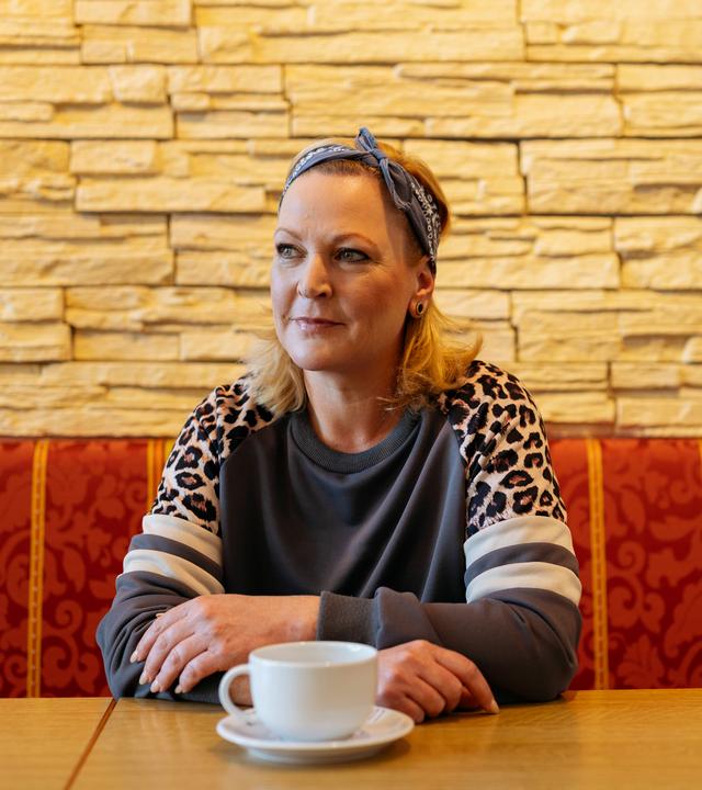 Martina sitzt auf einer Bank, an einem Tisch und schaut zur Seite. Sie hat die Hände auf den Tisch gelegt und verschränkt. Vor ihr steht eine weiße Tasse. 