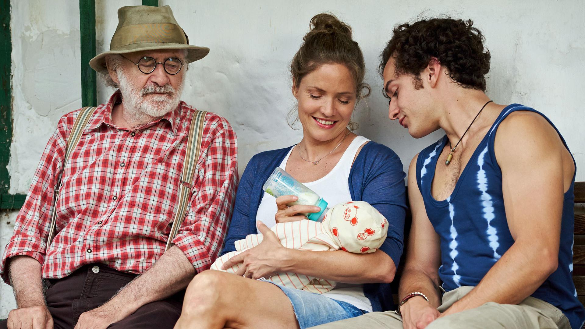 "Lena Lorenz - Mutter für drei Tage": Leo (Fred Stillkrauth) und Basti (Raban Bieling) sitzen links und rechts neben Lena (Patricia Aulitzky), die einen Säugling füttert und dabei lächelt.