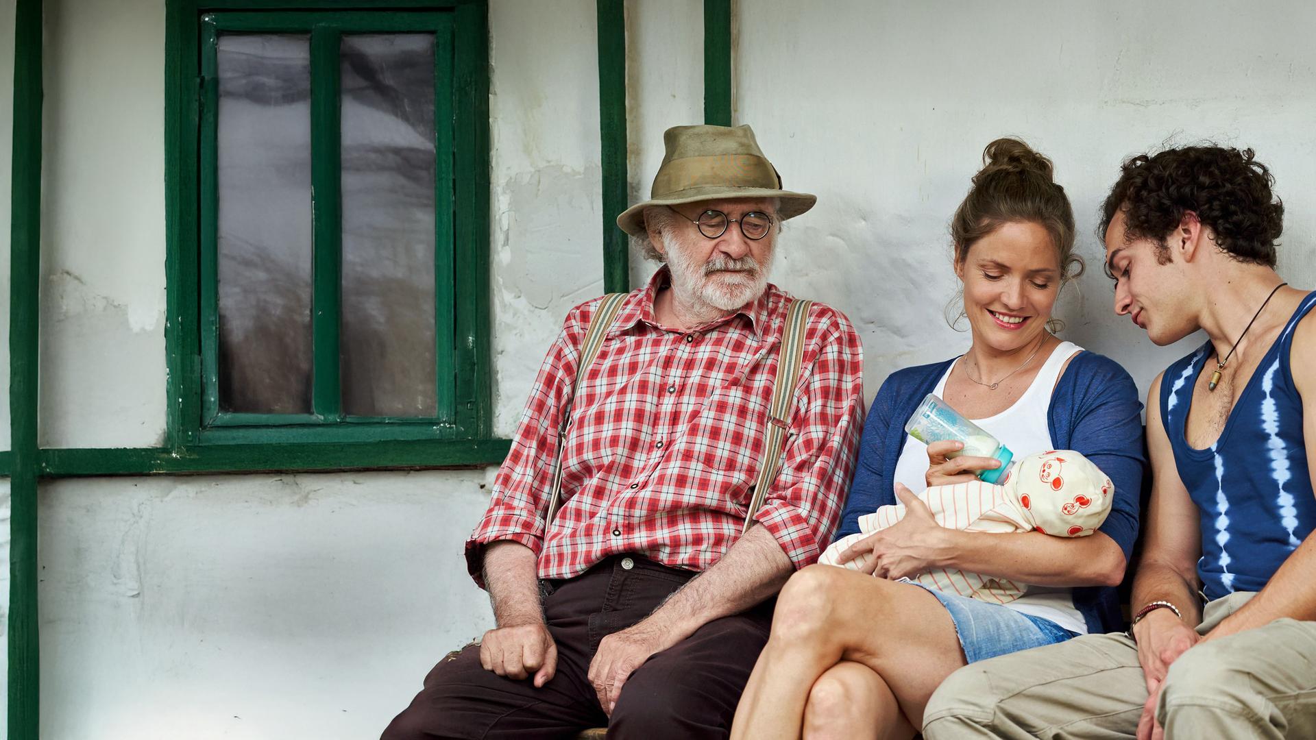 "Lena Lorenz - Mutter für drei Tage": Leo (Fred Stillkrauth) und Basti (Raban Bieling) sitzen links und rechts neben Lena (Patricia Aulitzky), die einen Säugling füttert und dabei lächelt.