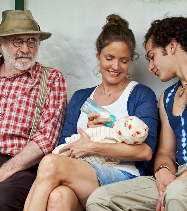 "Lena Lorenz - Mutter für drei Tage": Leo (Fred Stillkrauth) und Basti (Raban Bieling) sitzen links und rechts neben Lena (Patricia Aulitzky), die einen Säugling füttert und dabei lächelt.