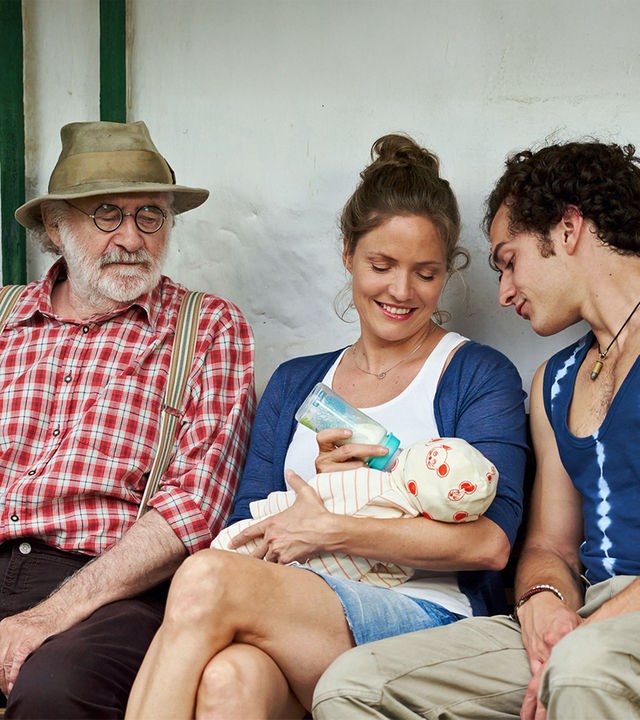 "Lena Lorenz - Mutter für drei Tage": Leo (Fred Stillkrauth) und Basti (Raban Bieling) sitzen links und rechts neben Lena (Patricia Aulitzky), die einen Säugling füttert und dabei lächelt.