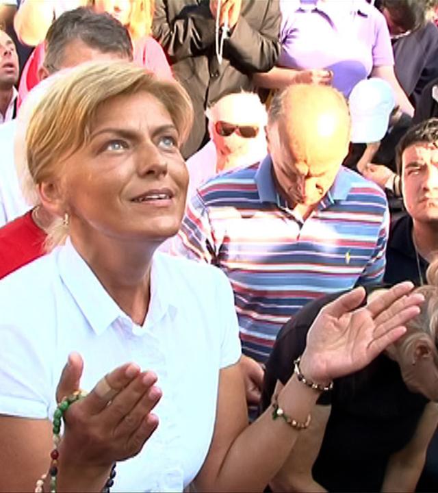 Das Bild zeigt eine Menschenmenge, die sich versammelt hat, um an einer spirituellen oder religiösen Veranstaltung teilzunehmen. Im Vordergrund ist eine Frau mit blonden Haaren zu sehen, die ein weißes Oberteil trägt. Sie hat ihre Hände geöffnet und zeigt eine gebetshaltung. Ihr Gesichtsausdruck ist konzentriert und andächtig.  Hinter ihr sind mehrere Personen sichtbar, die ebenfalls an der Veranstaltung teilnehmen. Einige blicken nach oben, während andere ihren Kopf gesenkt haben. Die Anwesenden haben verschiedene Gesichtsausdrücke, die von Ehrfurcht bis hin zu Ergriffenheit reichen. Es sind verschiedene Altersgruppen vertreten, einschließlich einer kleinen, weinenden Mädchen, das von einer Frau gehalten wird.  Die Umgebung wirkt lebhaft, und es sind viele Menschen in den Hintergrund zu sehen, die ebenfalls teilnehmen oder zuschauen. Die Kleidung variiert, wodurch ein vielfältiges und buntes Publikum entsteht. Die Veranstaltung scheint in einem Außenbereich stattzufinden, möglicherweise an einem religiösen oder spirituellen Ort.