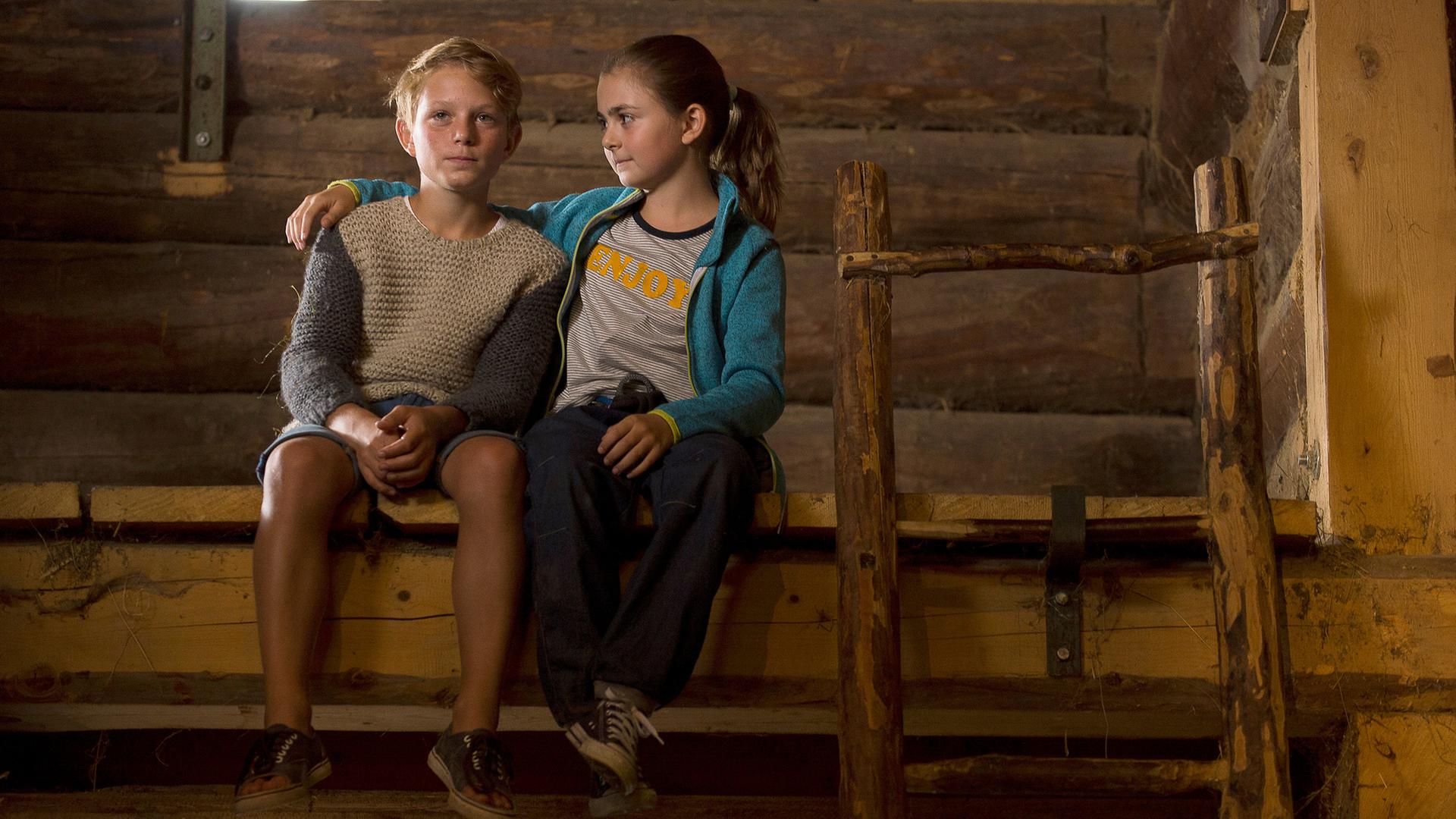 "Die Bergretter: Mutterliebe": Max (Niko Friesecke) und Mia (Mia-Sophie Ballauf) sitzen auf einem Holzvorsprung in einer Scheune. Max blickt sehr traurig. Mia hat ihren Arm um seine Schulter gelegt und blickt ihn an.