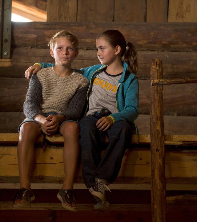 "Die Bergretter: Mutterliebe": Max (Niko Friesecke) und Mia (Mia-Sophie Ballauf) sitzen auf einem Holzvorsprung in einer Scheune. Max blickt sehr traurig. Mia hat ihren Arm um seine Schulter gelegt und blickt ihn an.