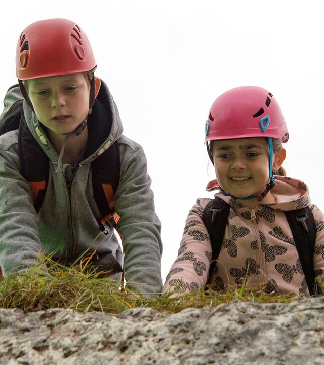 "Die Bergretter: Mutterliebe": Unterwegs in den Bergen, mit Rucksäcken und Wanderhelmen auf dem Kopf, knien Max (Nico Friesecke) und Mia (Mia-Sophie Ballauf) auf dem Boden und schauen ängstlich über einen Felsvorsprung in die Tiefe.