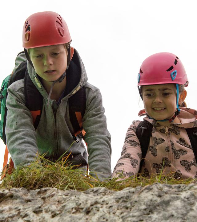 "Die Bergretter: Mutterliebe": Unterwegs in den Bergen, mit Rucksäcken und Wanderhelmen auf dem Kopf, knien Max (Nico Friesecke) und Mia (Mia-Sophie Ballauf) auf dem Boden und schauen ängstlich über einen Felsvorsprung in die Tiefe.