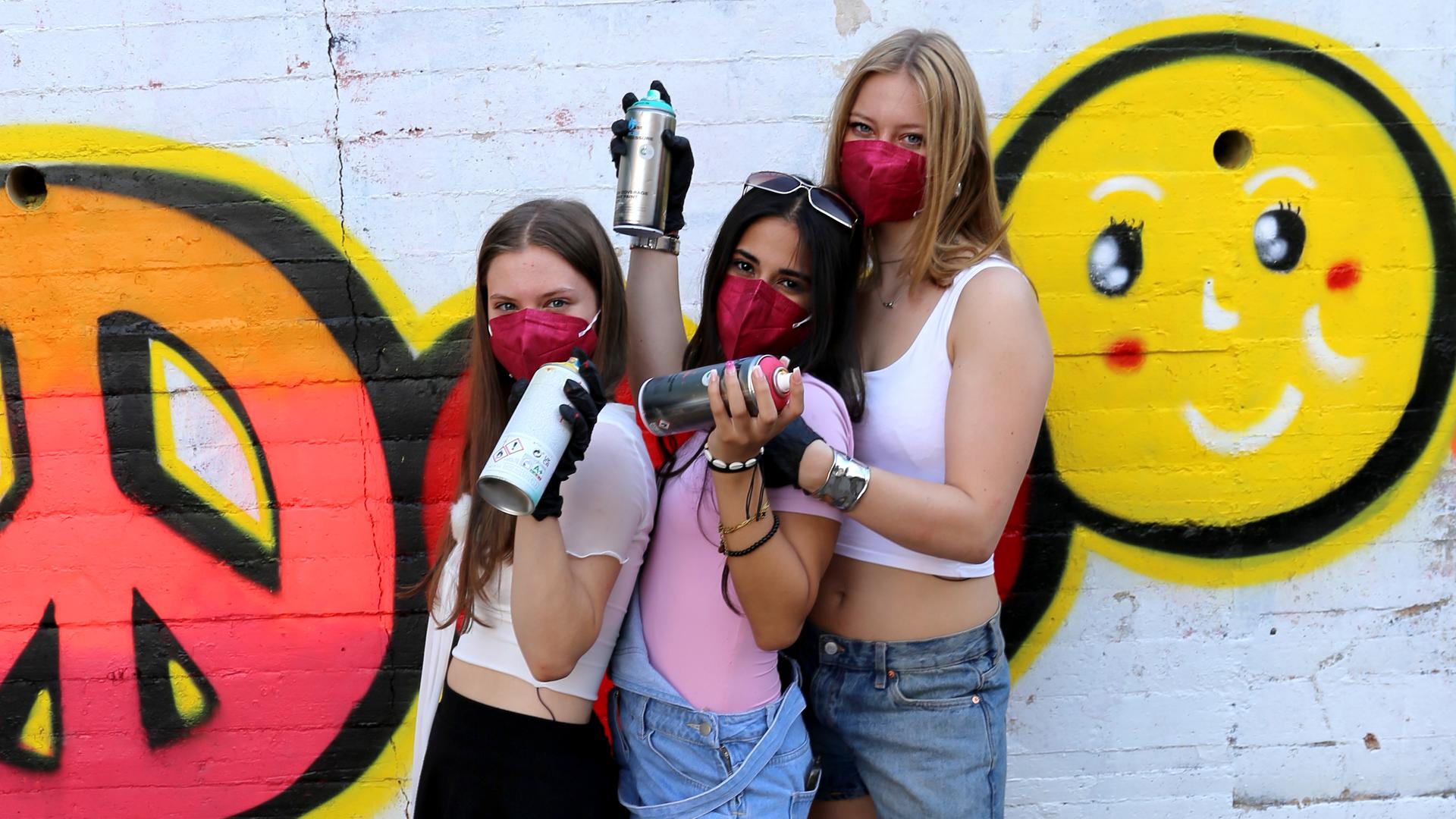 Josefine, Mahak und Johanna stehen vor einer Spraying-Wand und haben Sprühdosen in der Hand. Sie tragen jeweils eine Atemmaske.