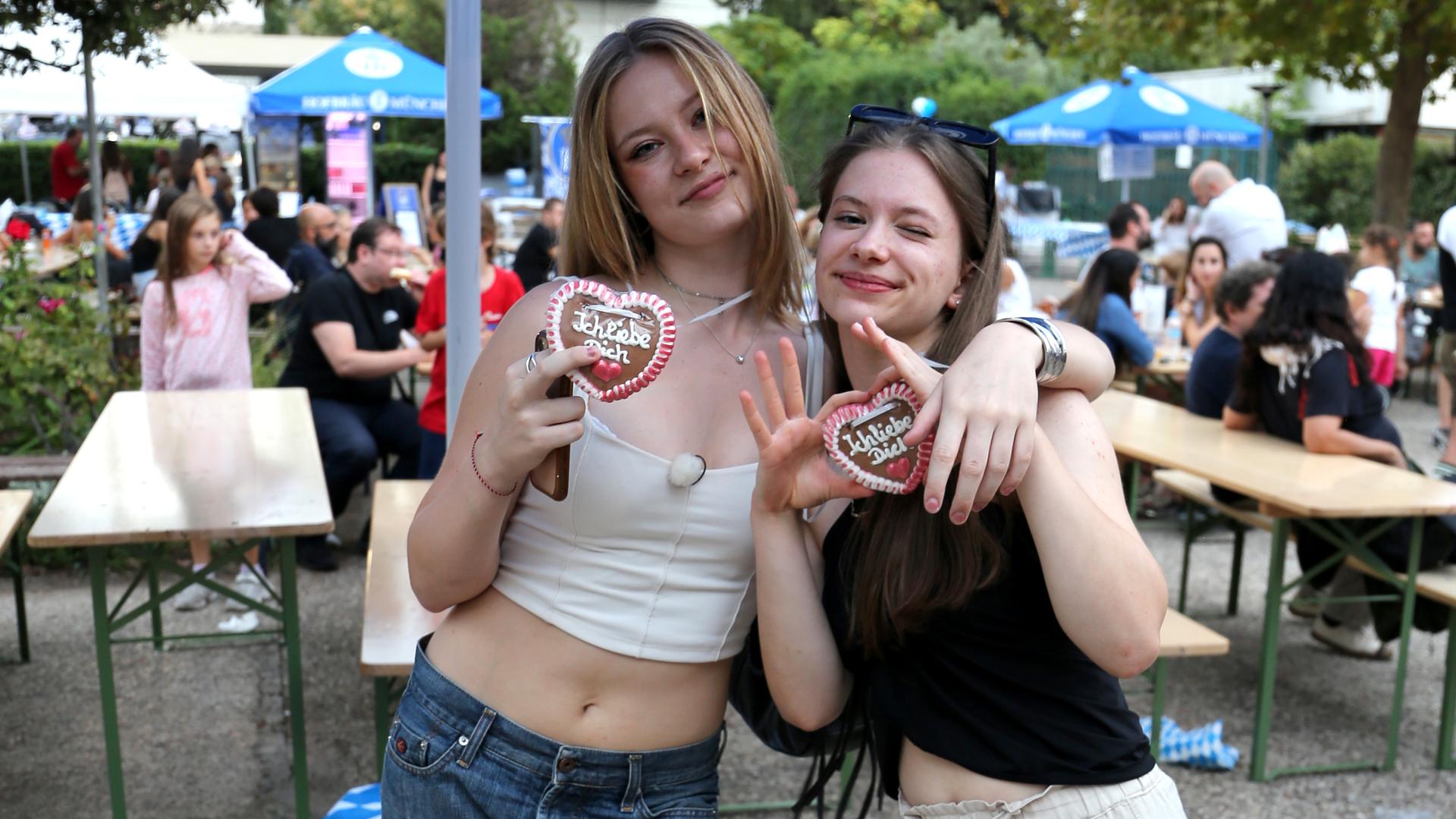 Johanna und Josefine stehen vor Holztischen auf einem Oktoberfest. Beide haben ein Lebkuchenherz um den Hals.