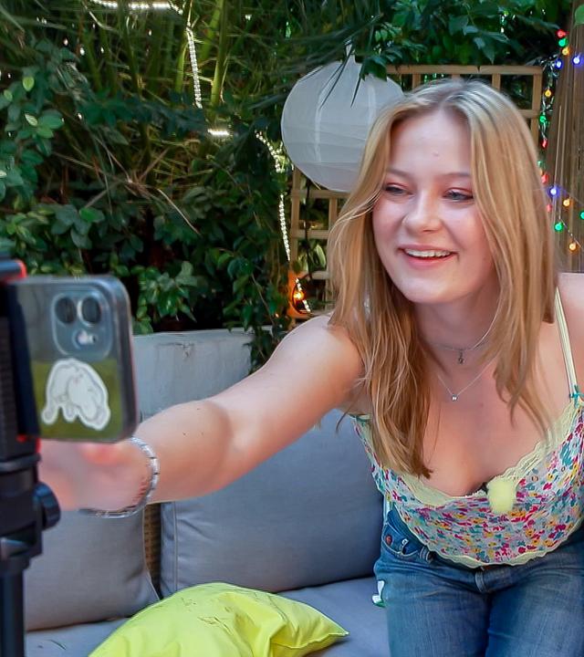 Johanna und Josefine sitzen auf einem Sofa. Johanna beugt sich zu einem Handy, welches vor ihnen auf einem Stativ steht, und startet die Aufnahme.