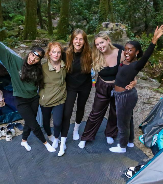 Fünf Mädchen stehen vor zwei Iglu-Zelten in einem Wald. Sie tragen Jogginghosen. Die Zelte sind offen, es liegen Schlafsäcke und Luftmatratzen darin. Vor den Zelten liegen die Turnschuhe der Mädchen. Die Fünf haben die Arme umeinander gelegt und lachen in die Kamera.
