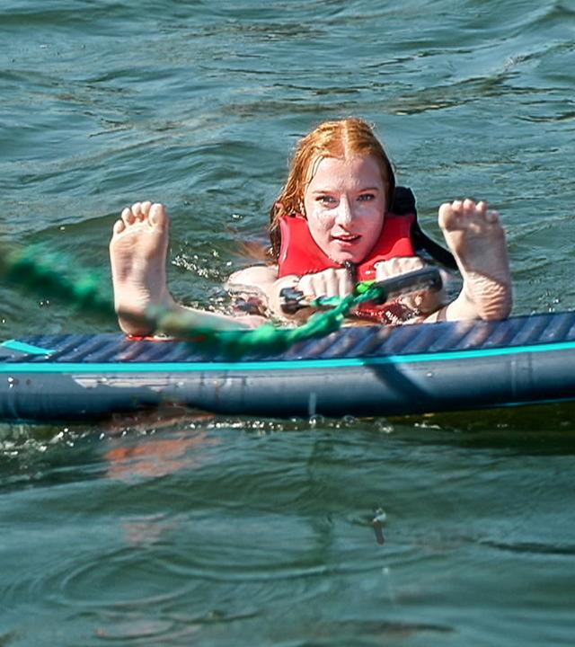 Mitten im Waser liegt ein Waveboard. Dahinter ist der Kopf eine rothaarigen Mädchens zu sehen, sie hält in der Hand eine grüne Zugleine. Die Füße streckt sie nach vorne in die Kamera, die Fersen liegen an der Kante des Boards auf.