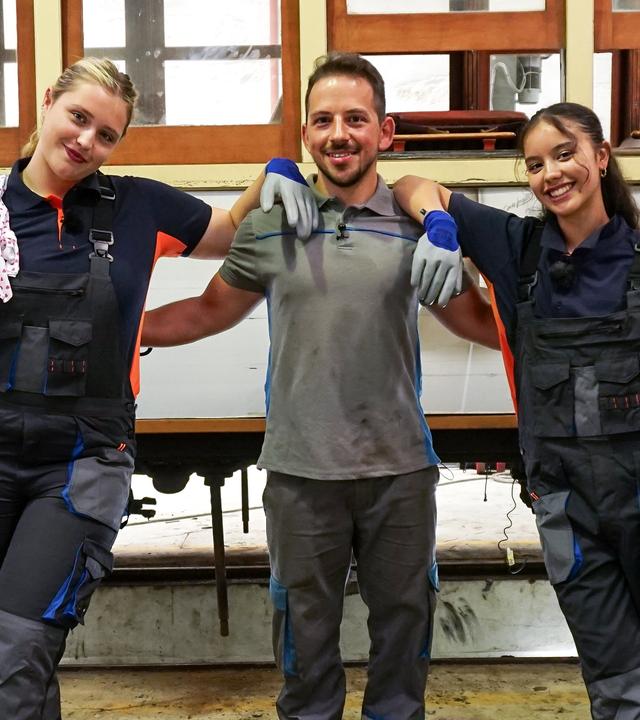 Zwei Mädchen haben sich auf die Schultern eines jüngeren Mannes gestützt, er legt freundschaftlich die Arme an ihre Rücken. Die Mädchen tragen blaue Arbeitsoveralls und grau-blaue Arbeitshandschuhe. Das dunkelhaarige Mädchen auf der rechten Seite des Fotos hält ein weißes Tuch mit roten Punkten locker in die Höhe, das blonde Mädchen auf der linken Seite hat sich so ein Tuch über die rechte Schulter geworfen. Im Hintergrund ist eine Trambahn zu sehen. Es handelt sich um eine Tram-Werkstatt in Porto, Portugal.