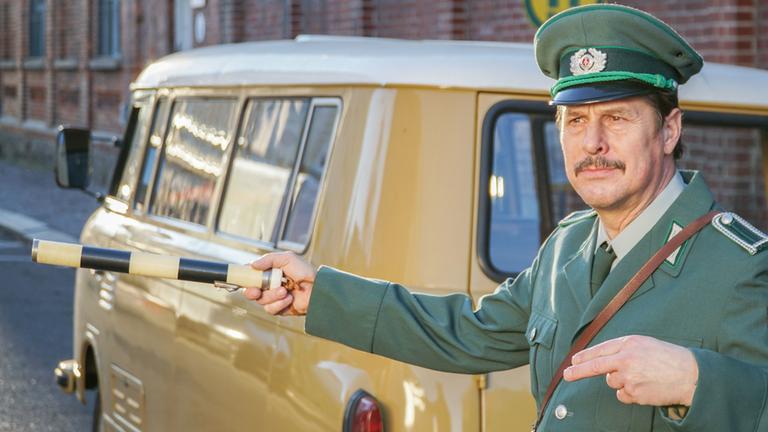 Ein DDR-Polizist steht in einer nachgestellten Szene auf der Straße und leitet den Verkehr um einen Tatort herum. Im Hintergrund steht ein gelber Transportwagen.