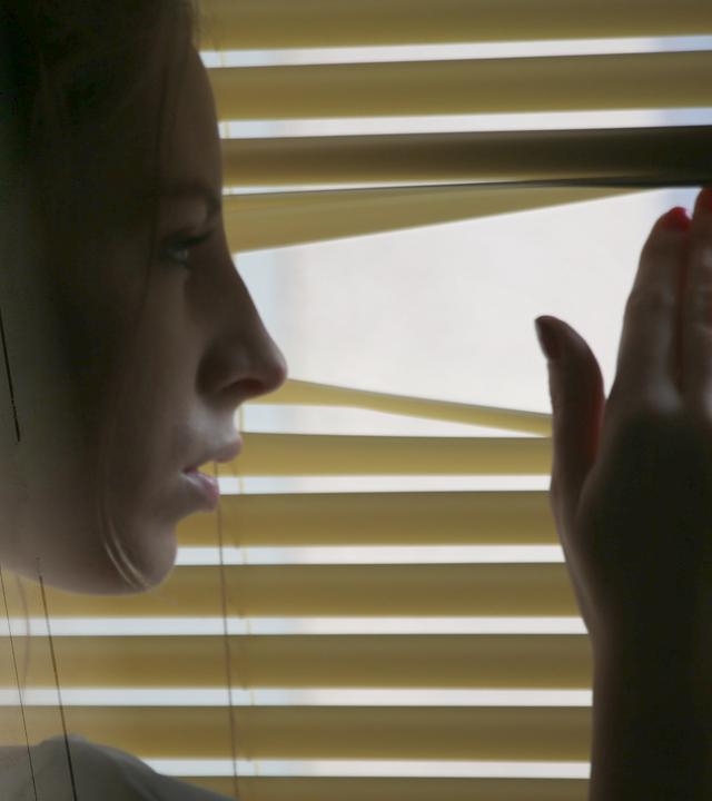 Eine Frau blickt verstohlen durch die Jalosien nach draußen.
