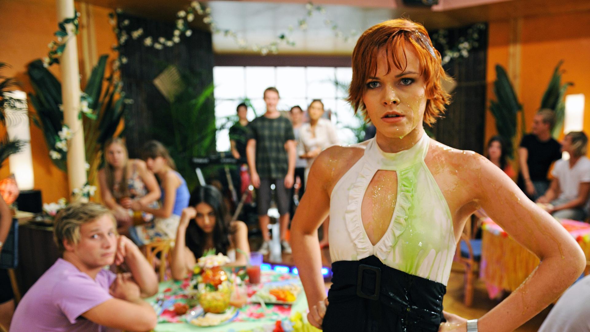 Sophie steht im Cafe und hat mit wütendem Blick die Fäuste in die Hüften gestemmt. Ihr Kleid ist mit grünem Saft verschmiert. Im Hintergrund sitzen Menschen an Tischen und schauen in ihre Richtung.