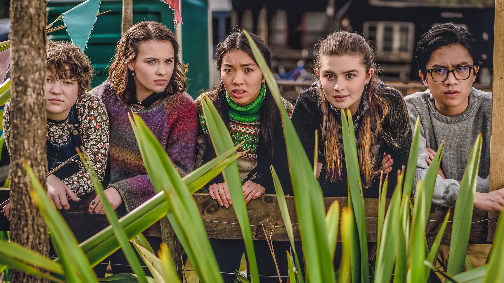 Draußen auf einem Fest hocken Stella, Issie, Caroline, Natascha und Caleb nebeneinander und schauen erwartungsvoll über eine grüne Pflanze nach vorn.