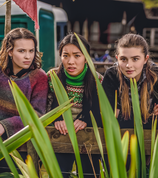 Draußen auf einem Fest hocken Stella, Issie, Caroline, Natascha und Caleb nebeneinander und schauen erwartungsvoll über eine grüne Pflanze nach vorn.