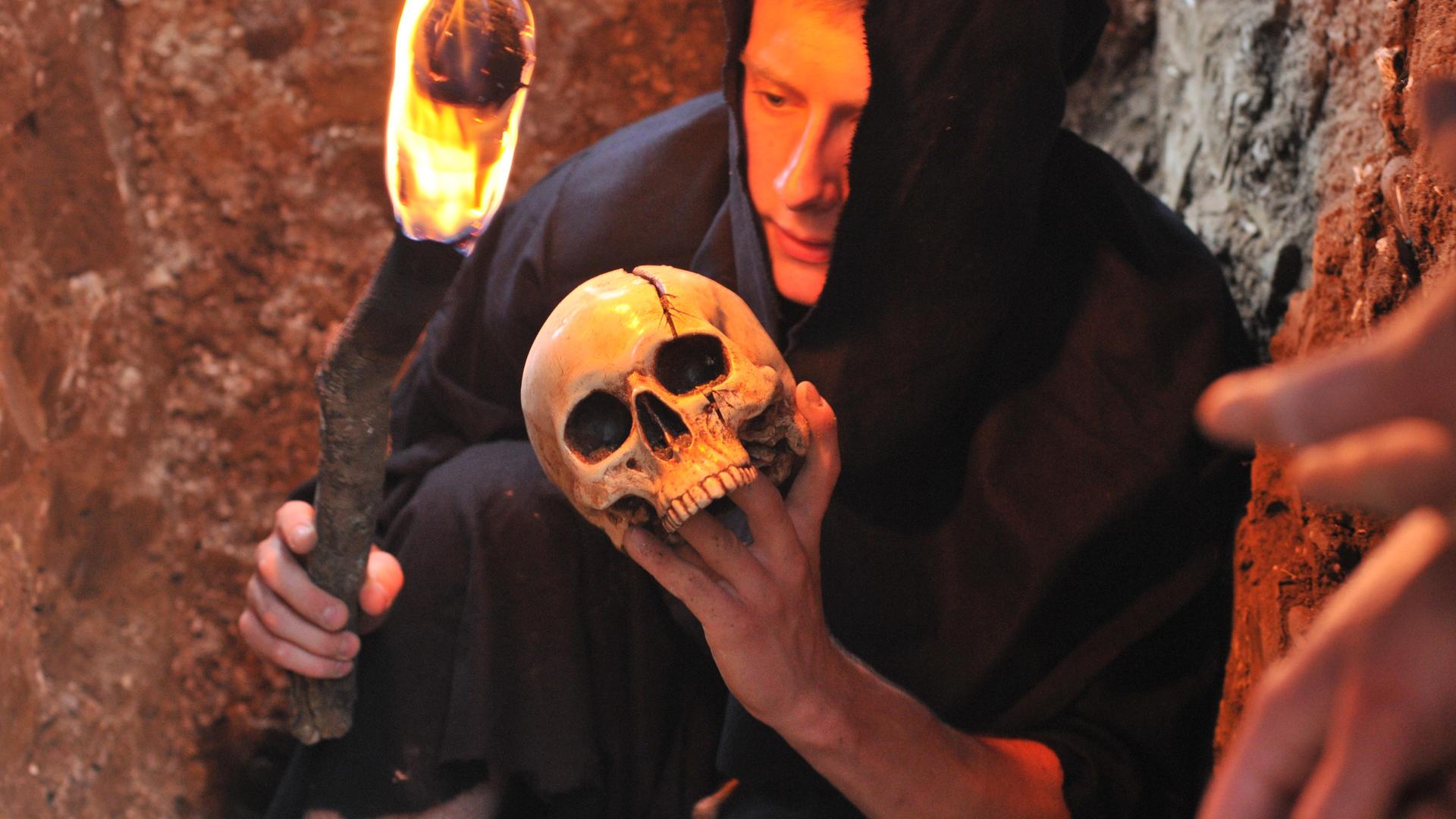 Ein Mann in schwarzer Kutte und Kapuze kniet auf dem Boden. Er hält in der einen Hand eine Fackel, in der anderen einen Totenschädel.