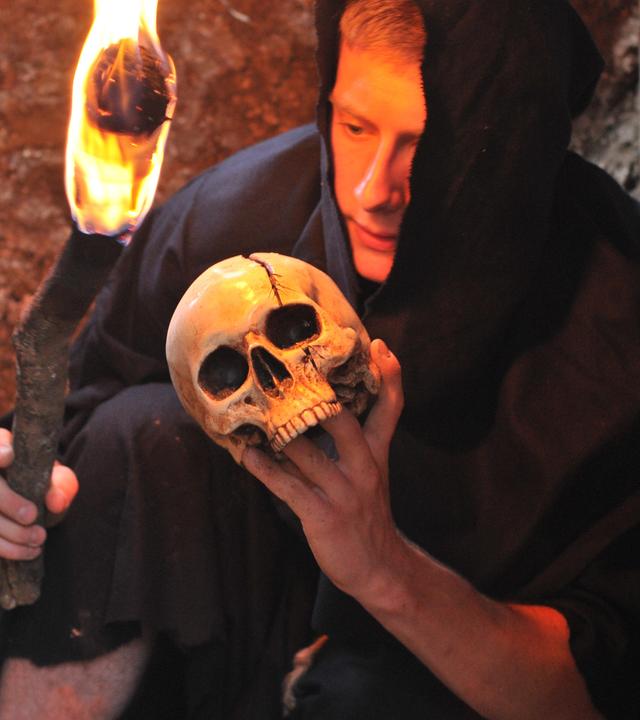 Ein Mann in schwarzer Kutte und Kapuze kniet auf dem Boden. Er hält in der einen Hand eine Fackel, in der anderen einen Totenschädel.