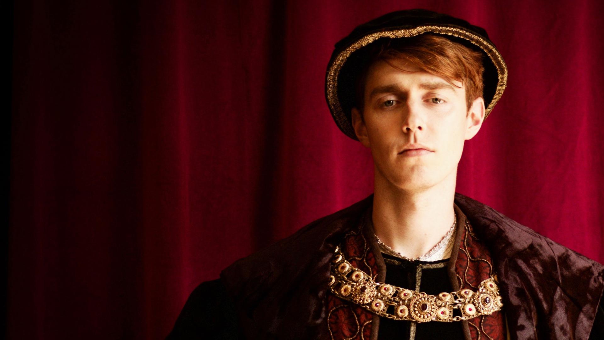 Spielszene: König Heinrich VIII. steht vor einem tiefroten Vorhang. Er hat rötliche Haare und trägt Hut und prachtvolle Kleidung des 16. Jahrhunderts in gedeckten Farben. Um den Hals trägt er eine große Goldkette.