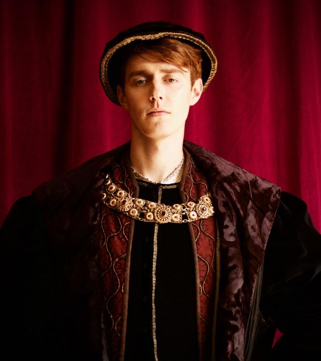 Spielszene: König Heinrich VIII. steht vor einem tiefroten Vorhang. Er hat rötliche Haare und trägt Hut und prachtvolle Kleidung des 16. Jahrhunderts in gedeckten Farben. Um den Hals trägt er eine große Goldkette.