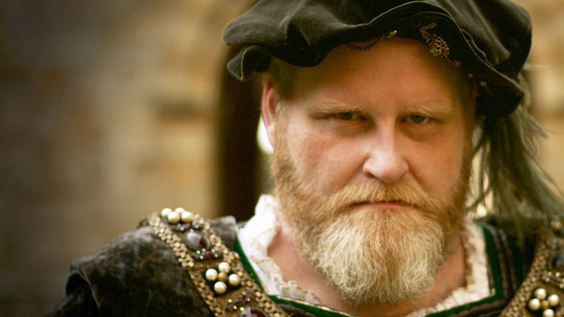 Spielszene. Porträt Heinrich VIII. mit rotblond graumeliertem Vollbart. Er trägt ein dunkelgrünes Samtbarett mit Feder und Brosche sowie einen dunklen Überrock im Renaissancestil.