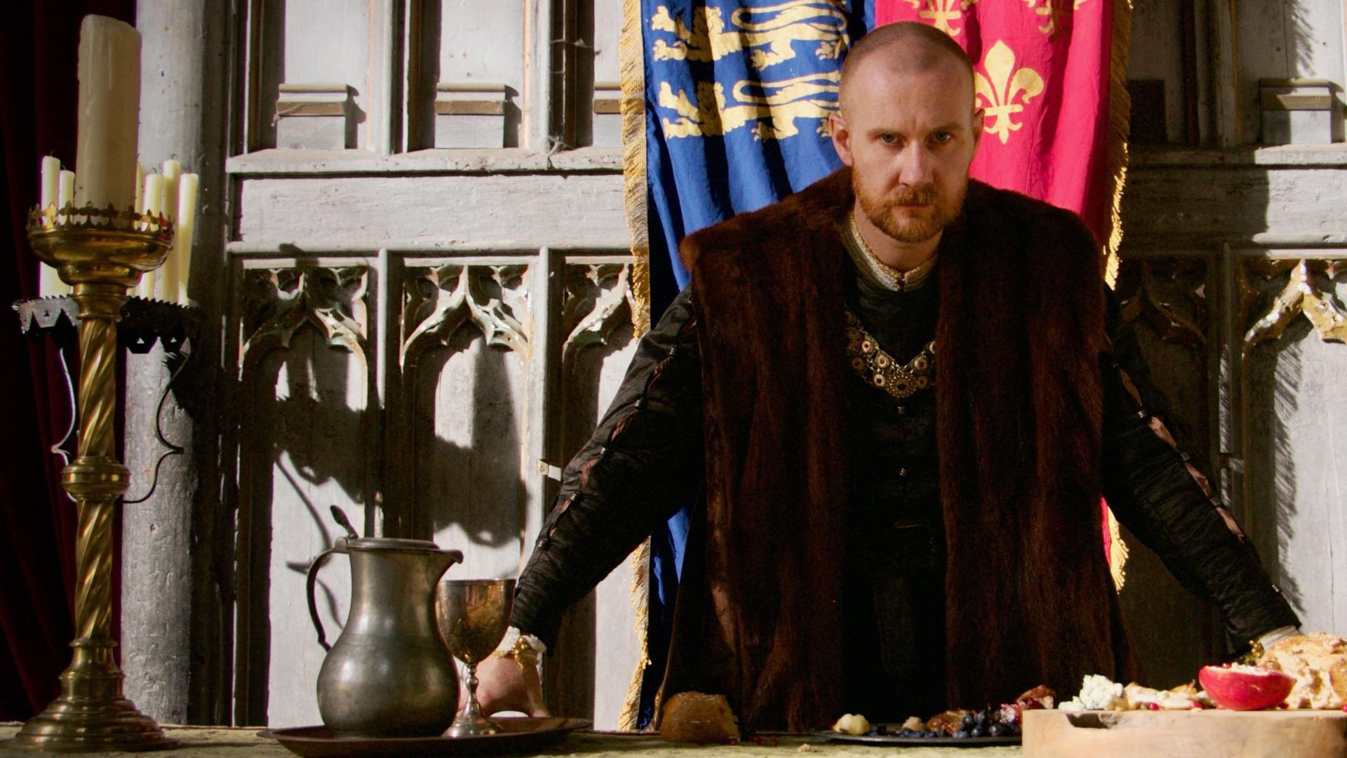 Spielszene: Hinter einem Tisch steht Heinrich VIII. mit kurzen, rötlichen Haaren, einem Bart und dunkler Kleidung. Seine Arme sind auf den Tisch gestützt, auf dem einrahmend zwei gewaltige goldene Kerzenleuchter sowie ein Krug und Essen stehen.