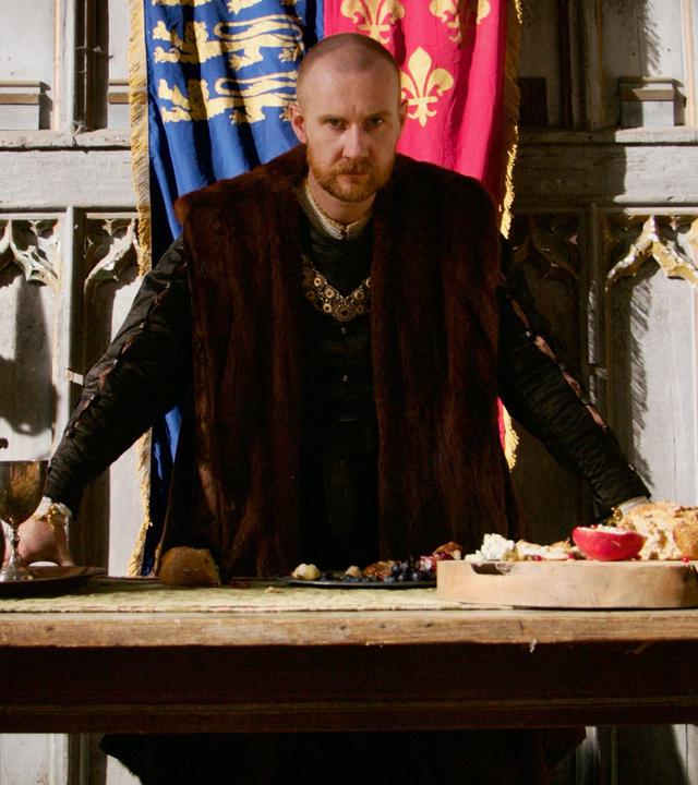 Spielszene: Hinter einem Tisch steht Heinrich VIII. mit kurzen, rötlichen Haaren, einem Bart und dunkler Kleidung. Seine Arme sind auf den Tisch gestützt, auf dem einrahmend zwei gewaltige goldene Kerzenleuchter sowie ein Krug und Essen stehen.