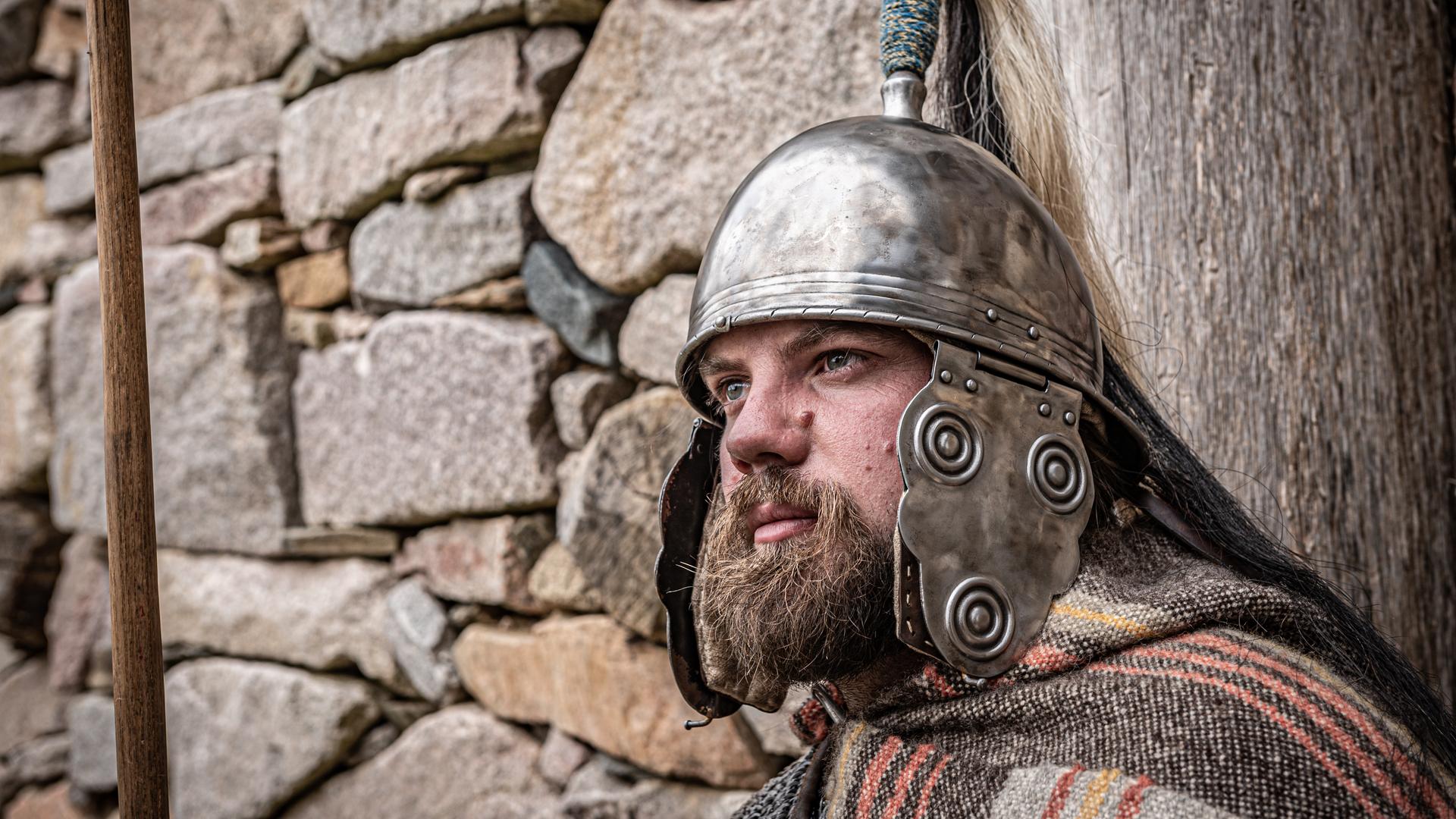 Ein bärtiger Mann in keltischer Rüstung lehnt sich mit starren Blick an eine Steinmauer.