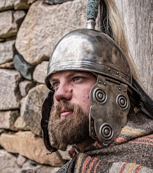 Ein bärtiger Mann in keltischer Rüstung lehnt sich mit starren Blick an eine Steinmauer.