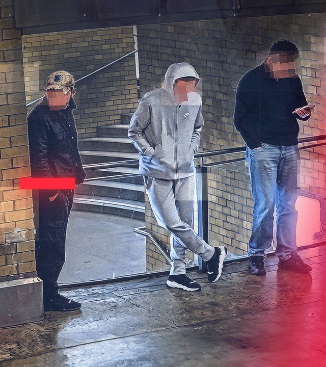 Drei junge Männer stehen in einem überdachten Bereich eines S- und U-Bahnhofes mit Backsteinwänden und Treppenaufgang. Die Gesichter sind verpixelt. Einer von ihnen trägt eine Tarnkappe und dunkle Kleidung, der Zweite einen grauen Trainingsanzug mit Kapuze, der Dritte einen dunklen Pullover und blickt auf ein Smartphone. Die Szene ist eine Aufnahme aus einer Überwachungskamera; rote grafische Overlay-Elemente und Rasterlinien sind über das Bild gelegt.