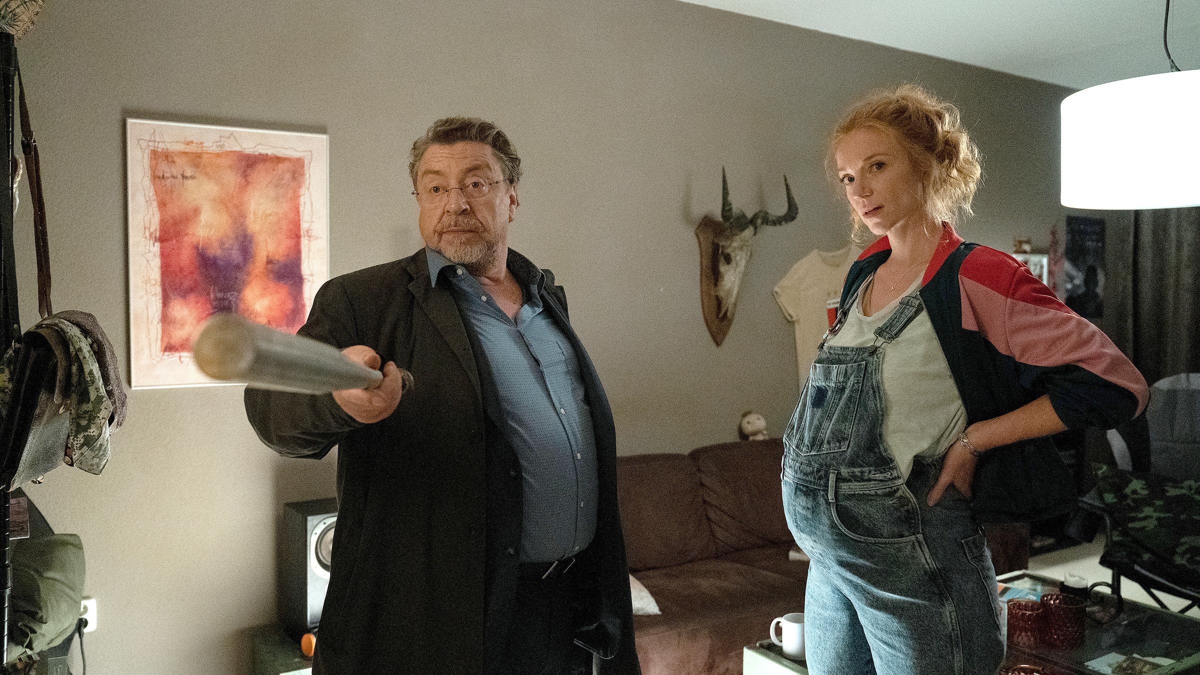 In einem Wohnzimmer: Erichsen (Armin Rohde) und die schwangere Mabel Kruse (Marleen Lohse) stehen nebeneinander. Erichsen hält einen Baseballschläger in der Hand.