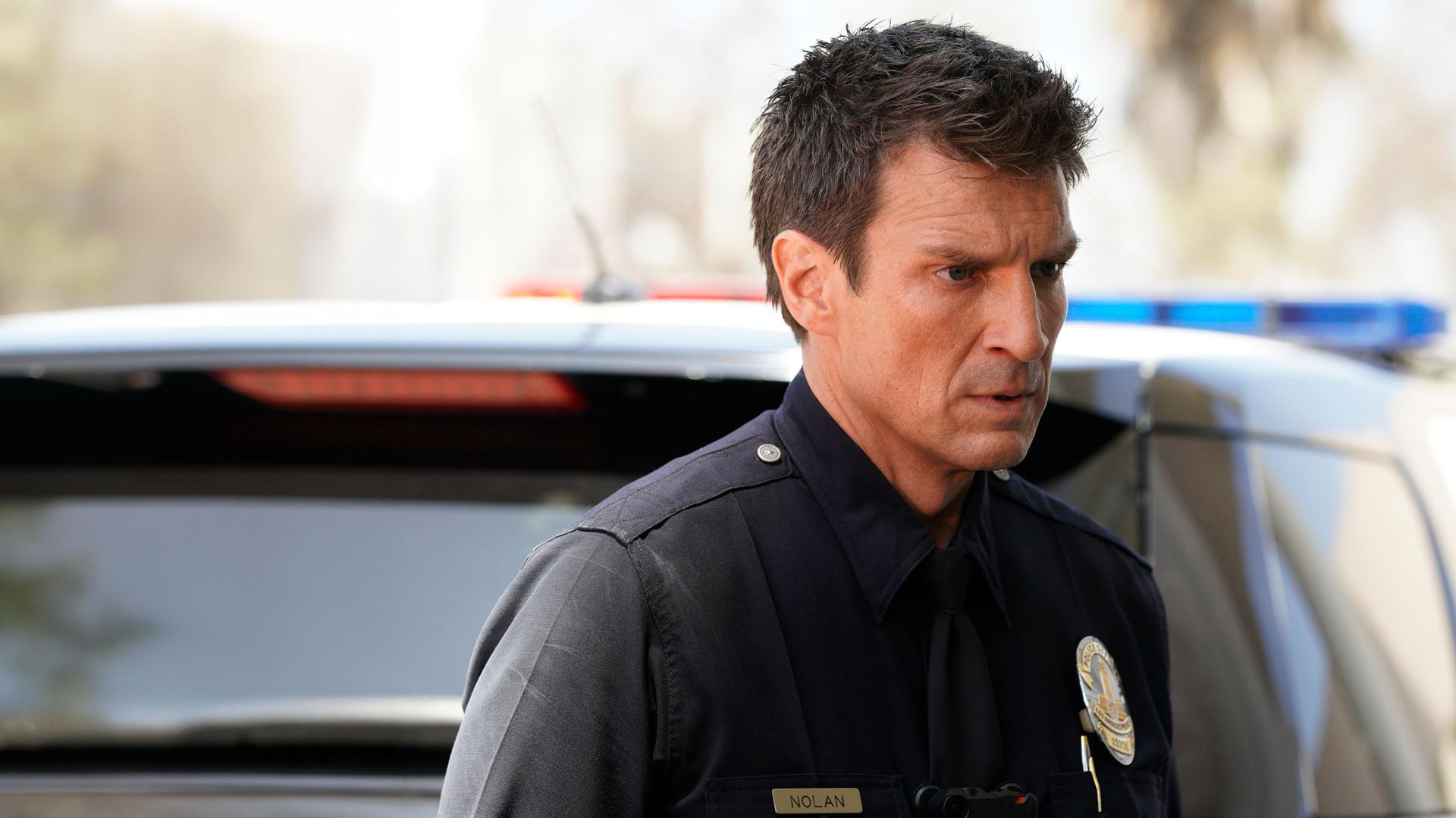 "The Rookie - Nachtschatten": John Nolan (Nathan Fillion) schaut mit entschlossenem Blick in die Ferne. Im Hintergrund ist ein Polizeiauto zu sehen.