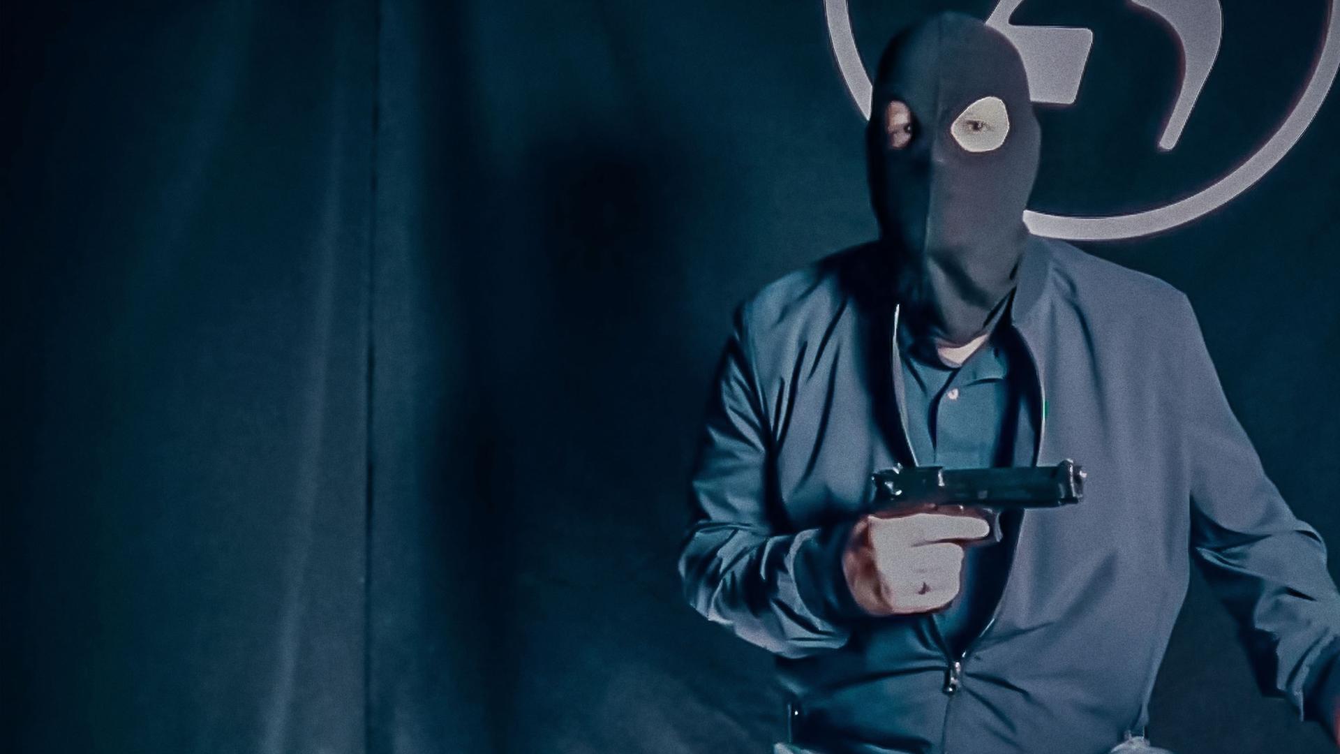 Ein Mann in einer Sturmmaske und mit einer Pistole in der Hand sitzt vor einem schwarzen Vorhang, auf dem die Zahl 25 aufgenäht ist. Das Bild sieht etwas verwaschen aus, als ob es aus einem Onlinevideo oder Livestream stammt.