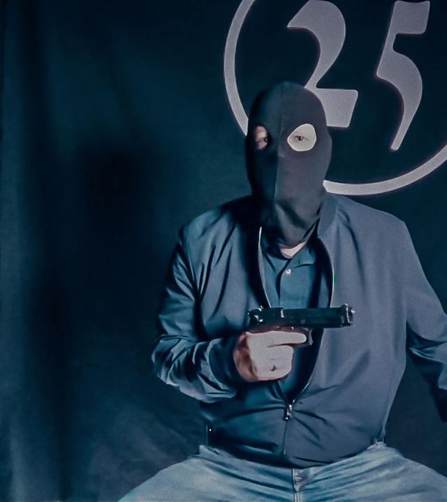 Ein Mann in einer Sturmmaske und mit einer Pistole in der Hand sitzt vor einem schwarzen Vorhang, auf dem die Zahl 25 aufgenäht ist. Das Bild sieht etwas verwaschen aus, als ob es aus einem Onlinevideo oder Livestream stammt.