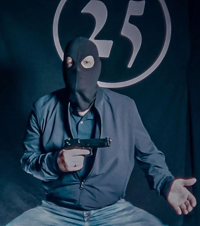 Ein Mann in einer Sturmmaske und mit einer Pistole in der Hand sitzt vor einem schwarzen Vorhang, auf dem die Zahl 25 aufgenäht ist. Das Bild sieht etwas verwaschen aus, als ob es aus einem Onlinevideo oder Livestream stammt.