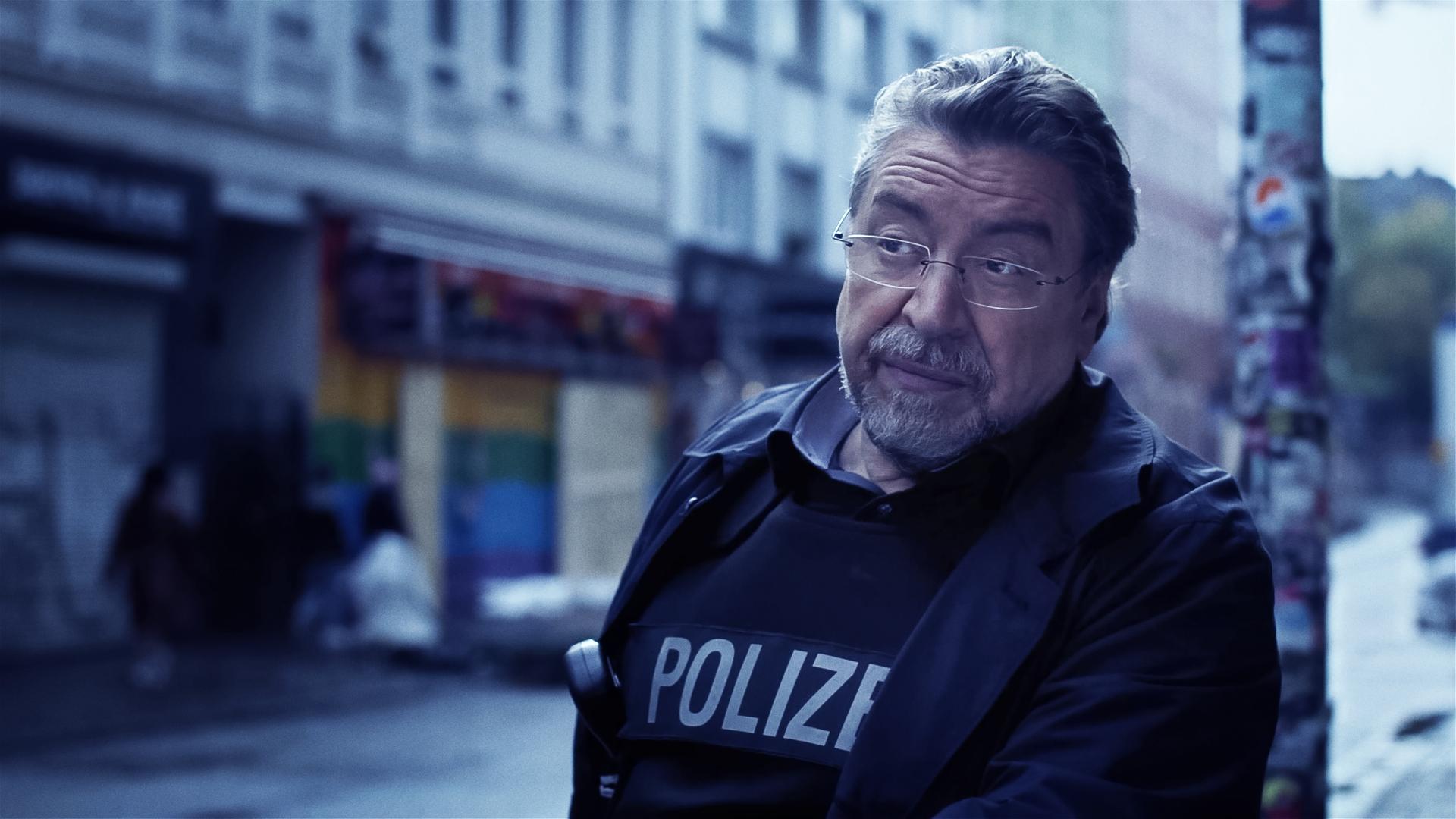 Ein Mann in Polizeiuniform (Armin Rohde) steht auf einer Straße und runzelt nachdenklich die Stirn. Im verschwommenen Hintergrund sieht man Häuserfassaden.