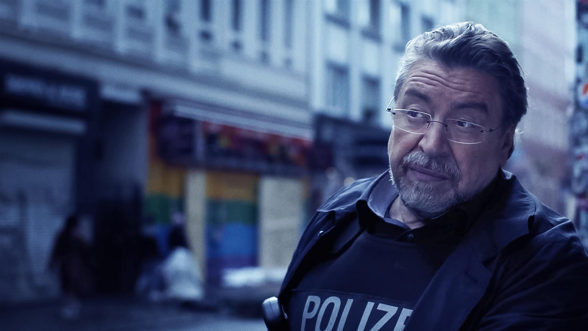 Ein Mann in Polizeiuniform (Armin Rohde) steht auf einer Straße und runzelt nachdenklich die Stirn. Im verschwommenen Hintergrund sieht man Häuserfassaden.